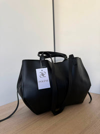 A black leather tote bag with dual top handles and a long shoulder strap, resting on a light wood surface against a plain off-white wall. A white paper tag with the brand name 'POLENE' is attached to the handle.